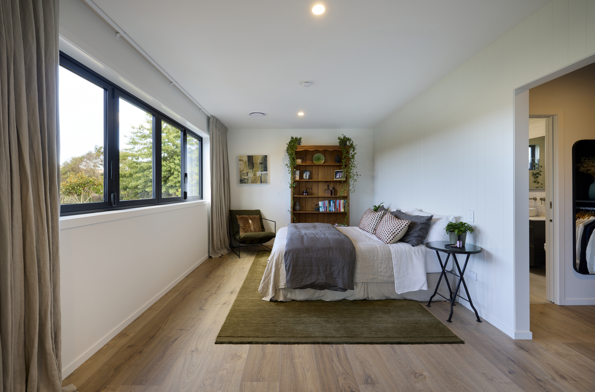 Spacious master bedroom in the Clevedon home with large black-framed windows overlooking greenery, timber floors, olive rug, timber bookshelf, and ensuite and walk-in wardrobe beyond.