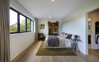 Spacious master bedroom in the Clevedon home with large black-framed windows overlooking greenery, timber floors, olive rug, timber bookshelf, and ensuite and walk-in wardrobe beyond.