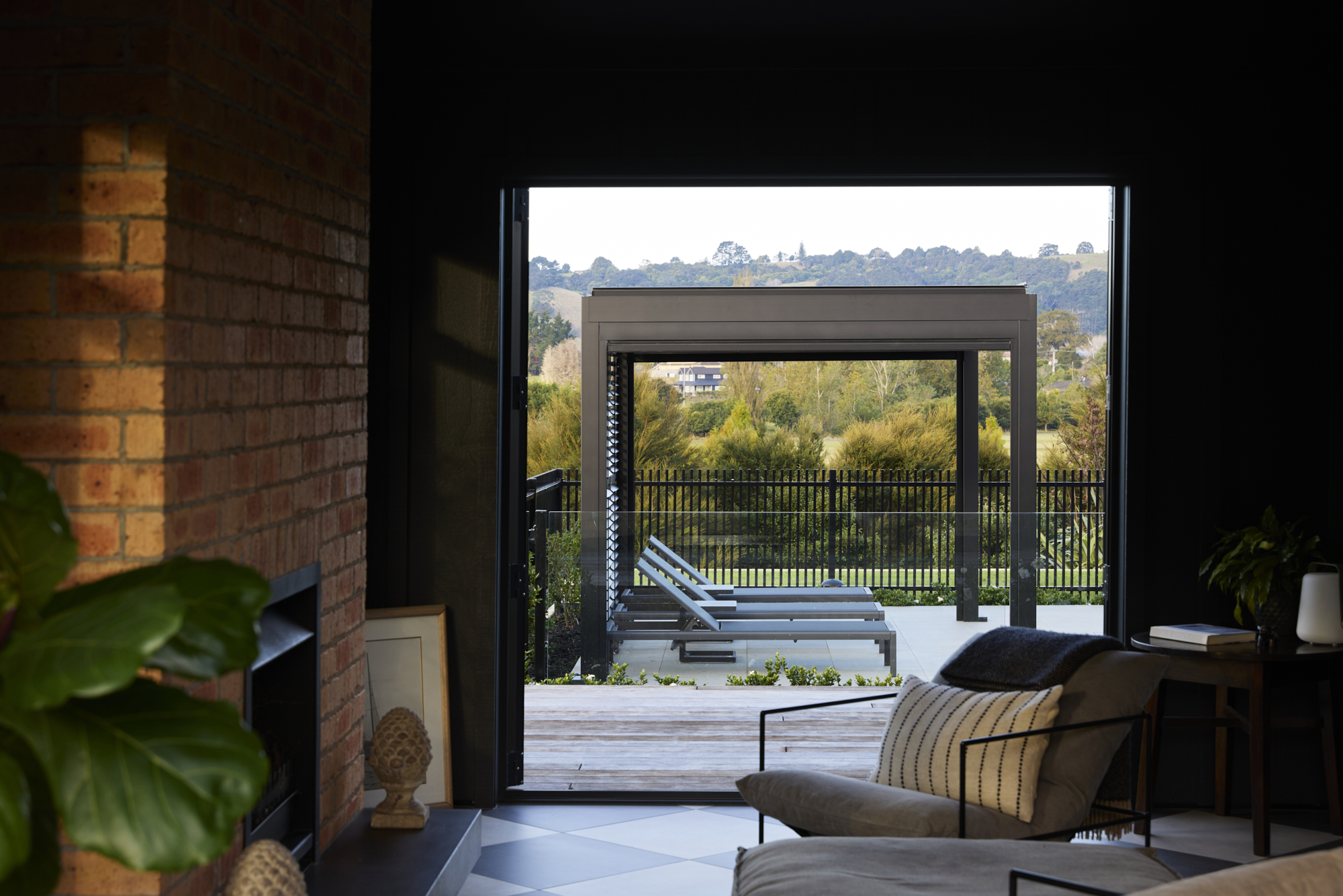Sunken lounge interior featuring a full-height exposed brick fireplace wall, dark ceiling, checkerboard flooring, woven pendant light, and open bifold doors framing garden views.