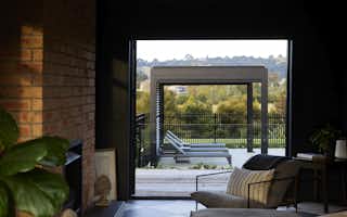 Sunken lounge interior featuring a full-height exposed brick fireplace wall, dark ceiling, checkerboard flooring, woven pendant light, and open bifold doors framing garden views.