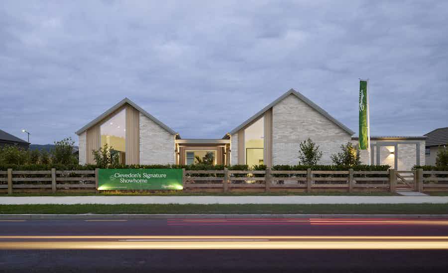 Street-facing exterior of the Clevedon Signature Showhome at twilight, showing the dual-gable white brick facade, timber-framed entry, and Signature Homes signage behind a timber post-and-rail fence.