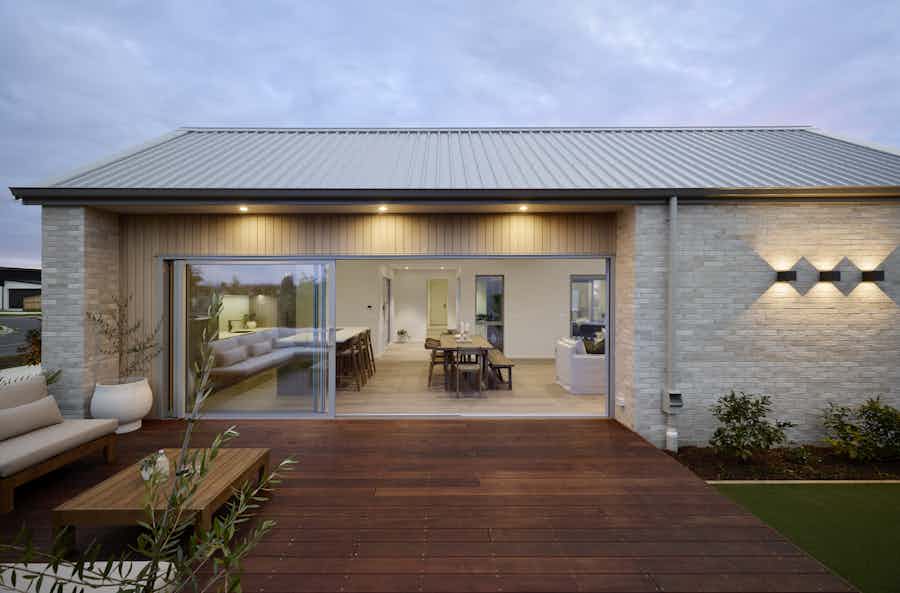 Rear view of the Clevedon Signature Showhome at dusk, with full-width stacking glass sliders open to a hardwood timber deck and outdoor seating area, revealing the open-plan dining and living interior.