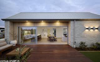 Rear view of the Clevedon Signature Showhome at dusk, with full-width stacking glass sliders open to a hardwood timber deck and outdoor seating area, revealing the open-plan dining and living interior.
