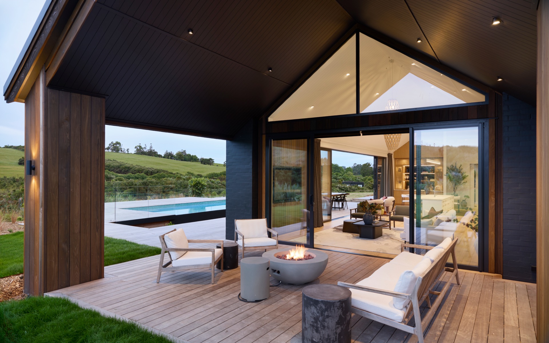Covered outdoor lounge with fire pit, timber decking, and views across the pool and rural landscape.