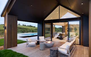 Covered outdoor lounge with fire pit, timber decking, and views across the pool and rural landscape.