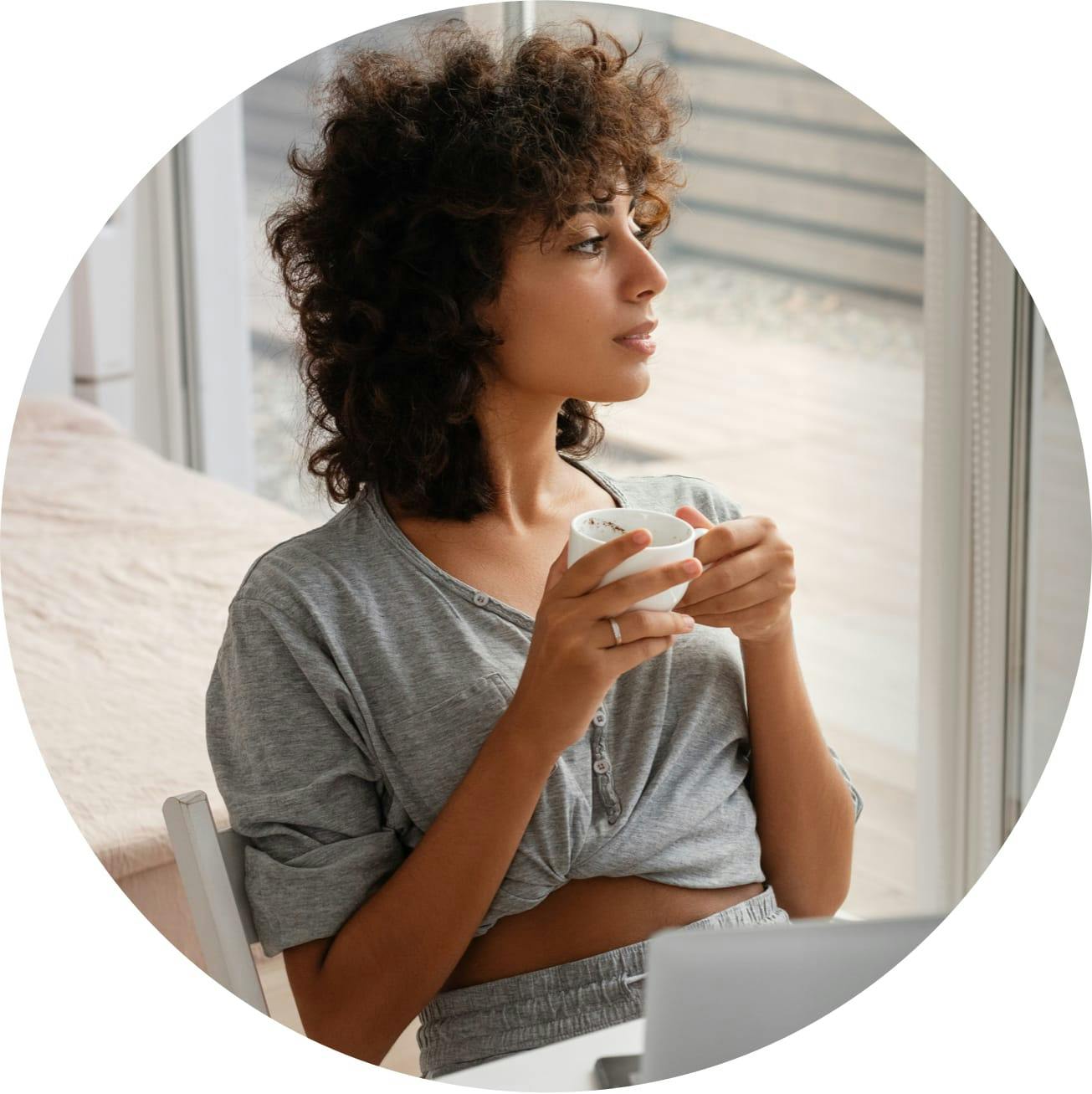Attractive woman in comfy clothes, holding a cup in two hands, looking off to her left.