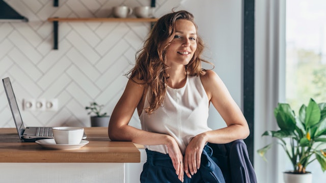 Female in her 20s with long wavy auburn hair sitting at a blonde wood and white kitchen benchtop with a laptop, cup and saucer