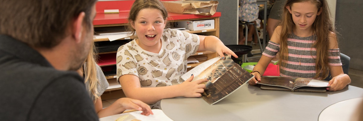 Girl in a classroom holds up book, pointing to a page