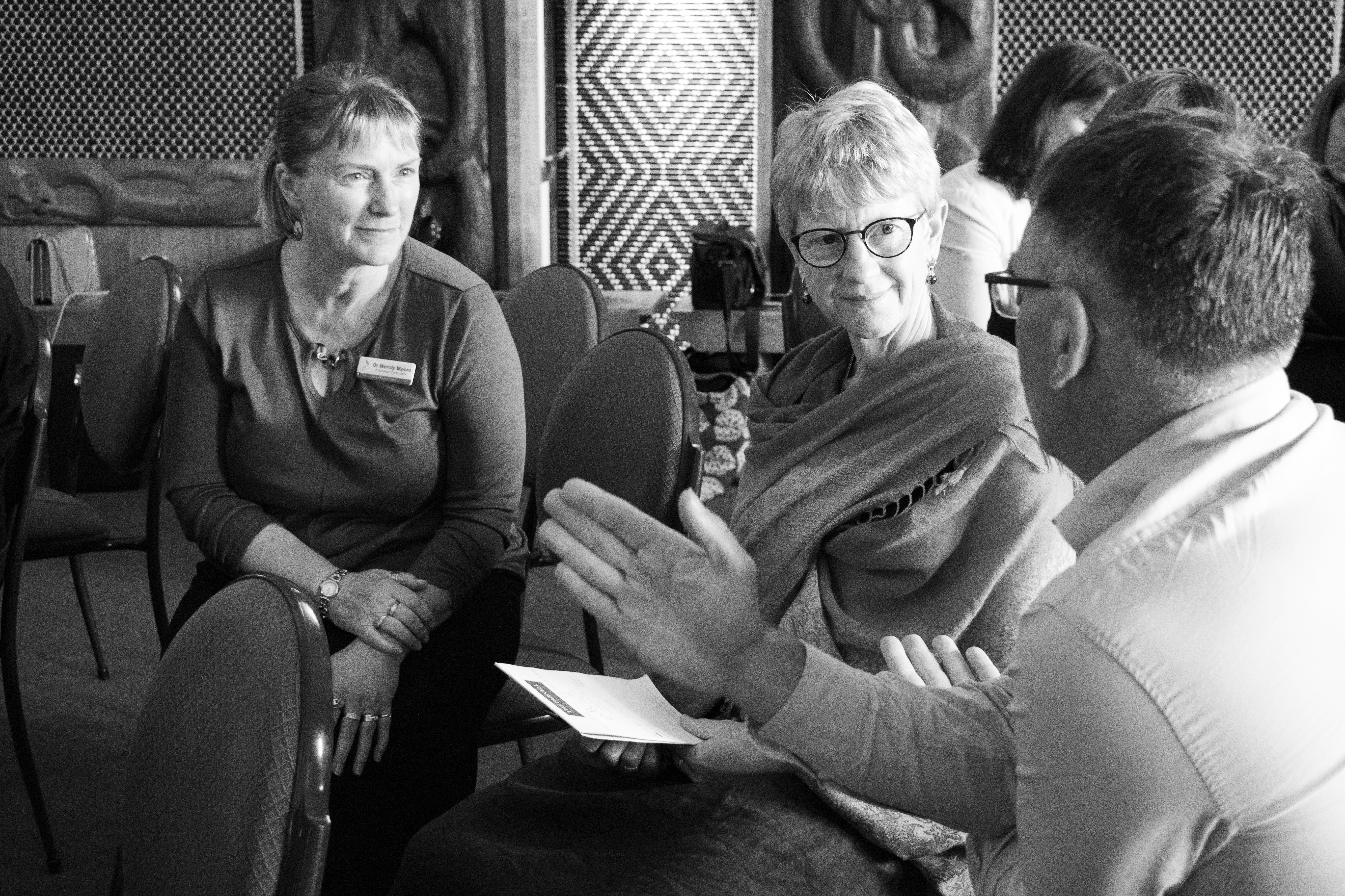 Three people sitting inside a whare (at marae). They are facing each other and appear to be in discussion