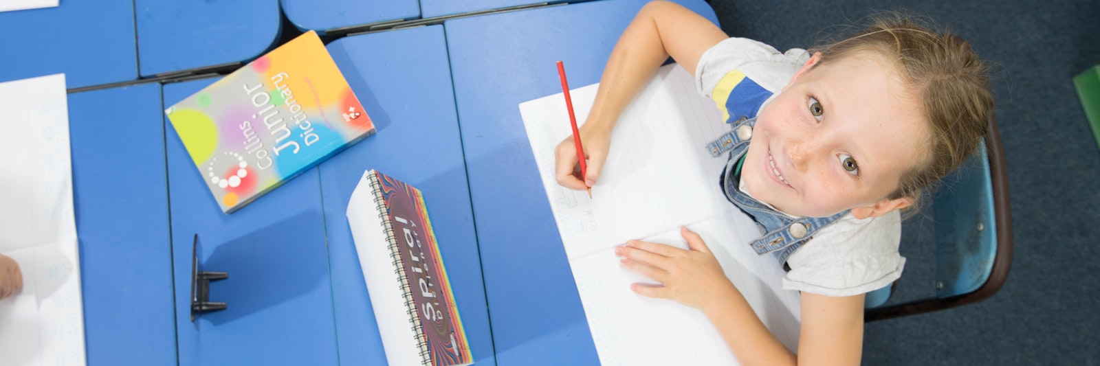 Child sitting at a desk with a dictionary, pen, and notebook. She looks at an overhead camera.