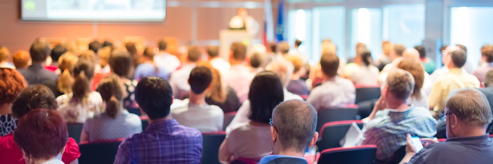 People sitting in a conference presentation, looking at a speaker and display screen