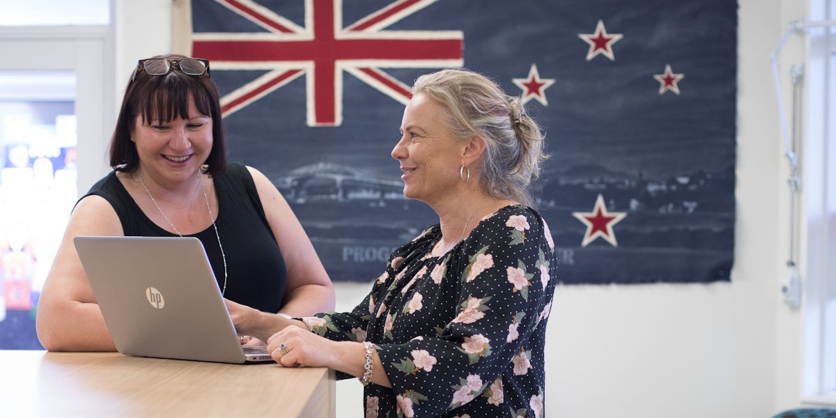 Two women standing in front of a New Zealand flag, looking at a computer and smiling