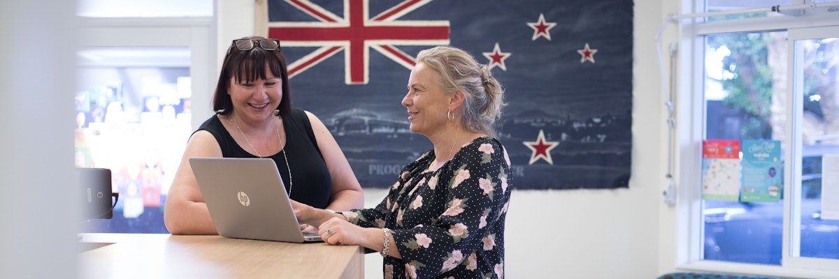 Two women standing in front of a New Zealand flag, looking at a computer and smiling