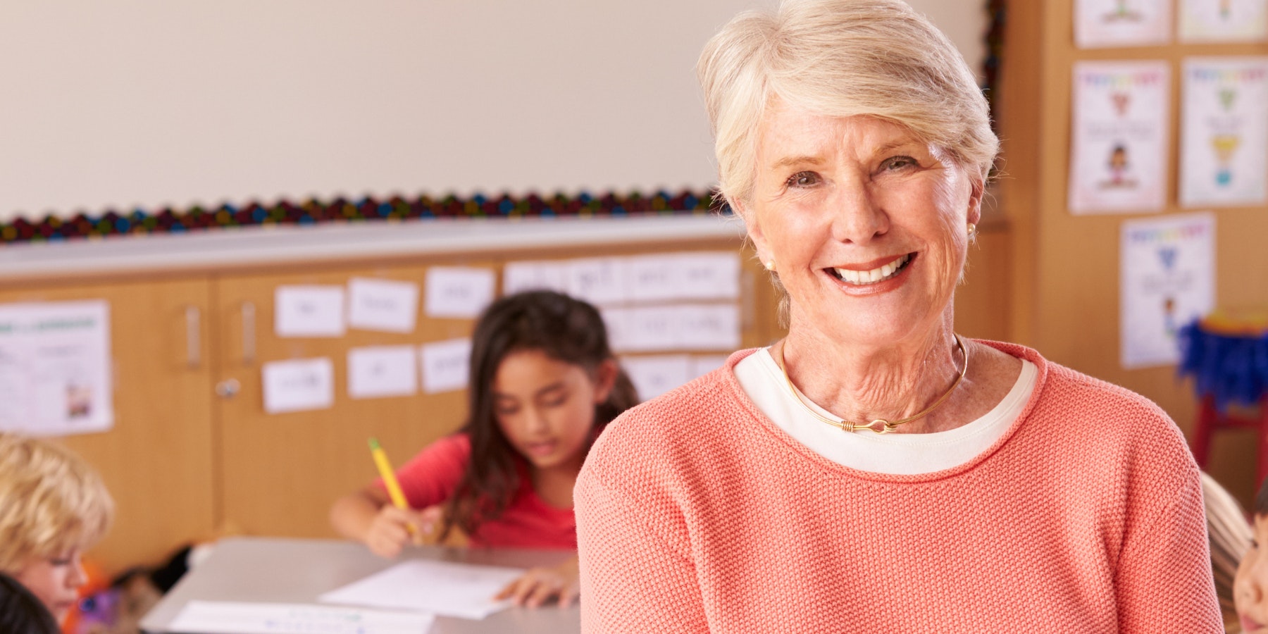 Teacher stands in a classroom with students in it. She is smiling at the camera.