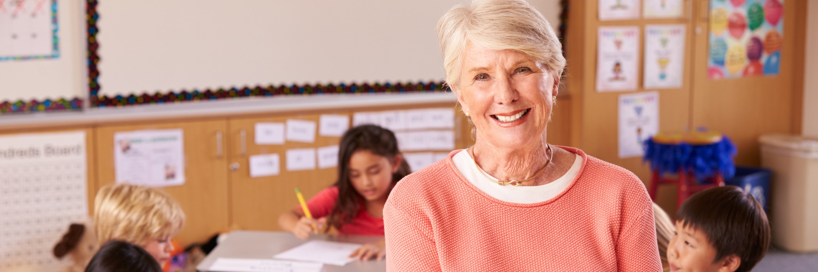 Teacher stands in a classroom with students in it. She is smiling at the camera.