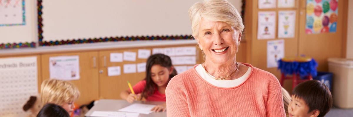 Teacher stands in a classroom with students in it. She is smiling at the camera.
