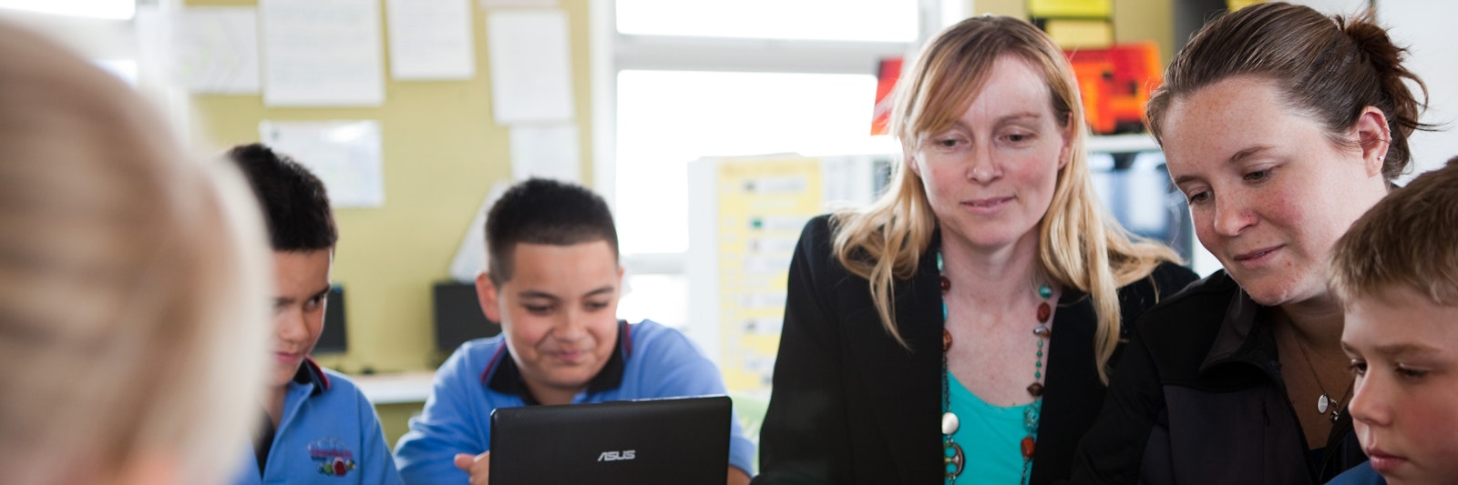 Two teachers sit in a classroom, surrounded by students