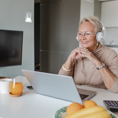 Woman sits at her kitchen bench. She is wearing headphones and looking at a computer screen