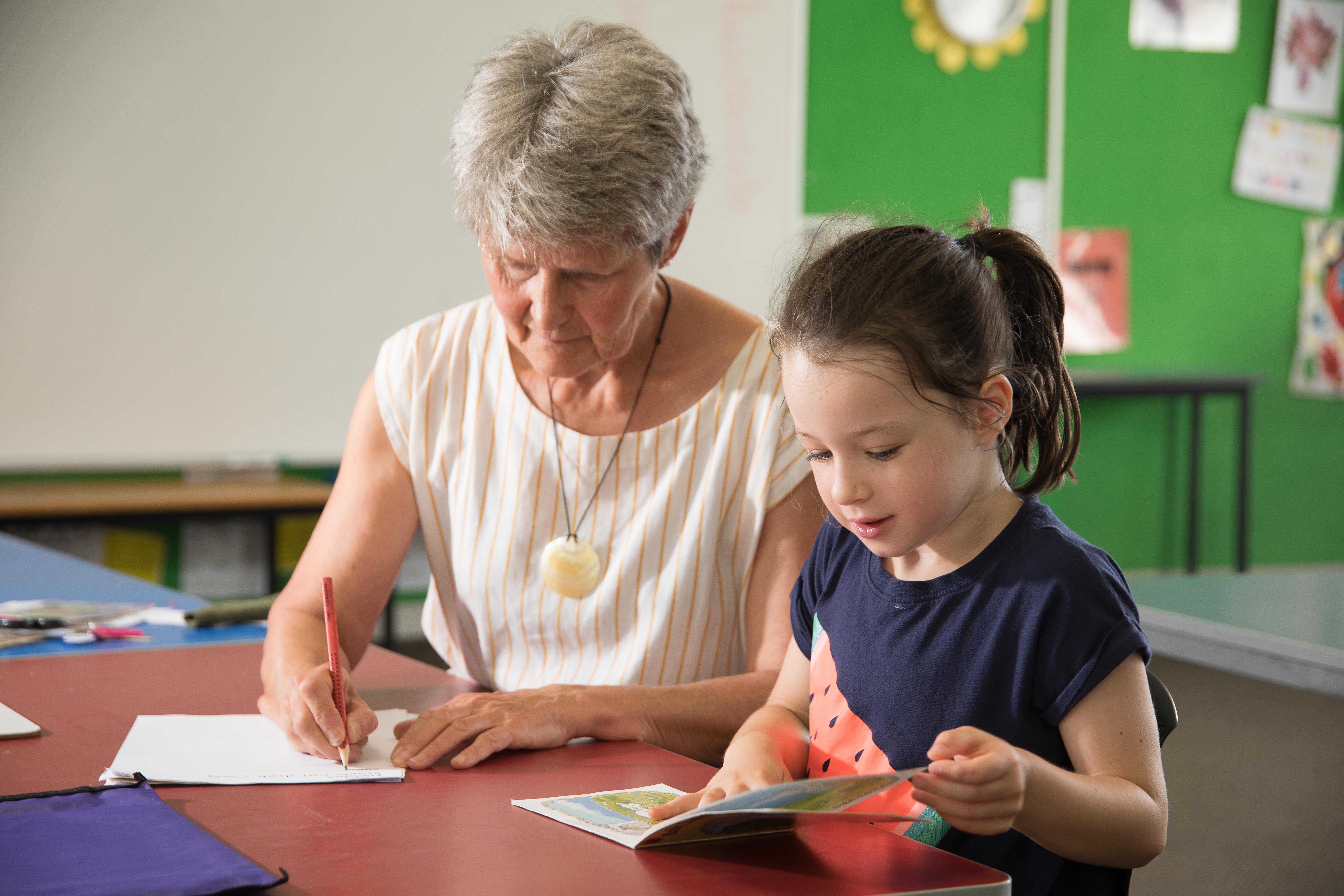 Female teacher sits beside a junior school student at a desk in a classroom. The teacher is writing on sheet of paper. The student is reading from a book.