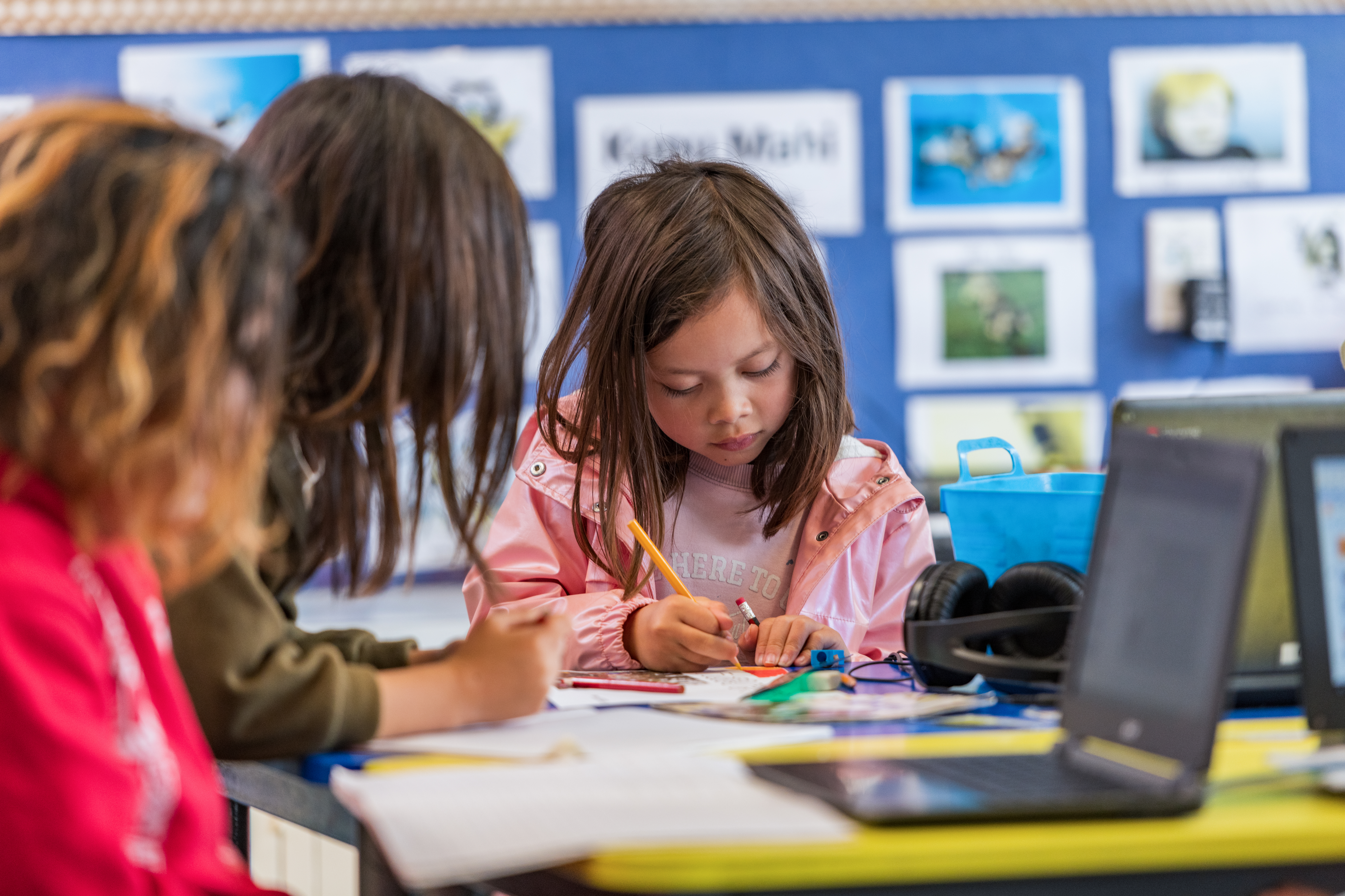 A female primary school student sits at a table in a typical New Zealand classroom. She is writing in a book with a pencil. Two students are sitting to her right. They both have their heads down. One is writing with the pencil. The other has an open laptop in front of them.