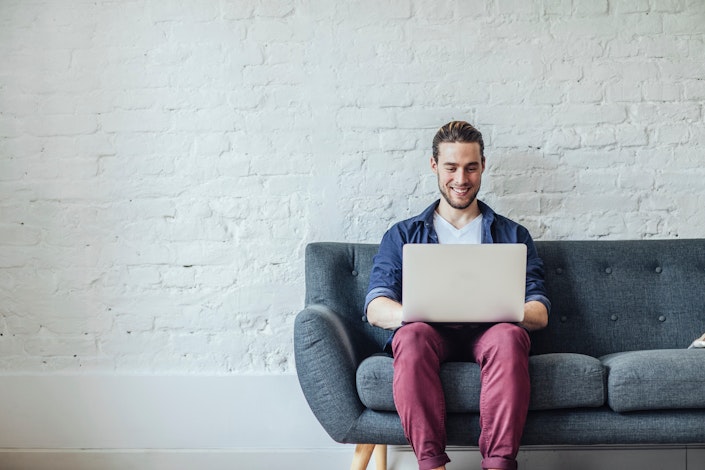 junger Mann sitz mit Laptop auf dem Schoß auf dem Sofa