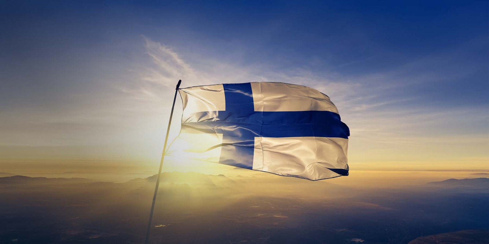 Finlands flagga vajar i solnedgång över ett landskap med berg och himmel.