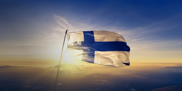 Finlands flagga vajar i solnedgång över ett landskap med berg och himmel.