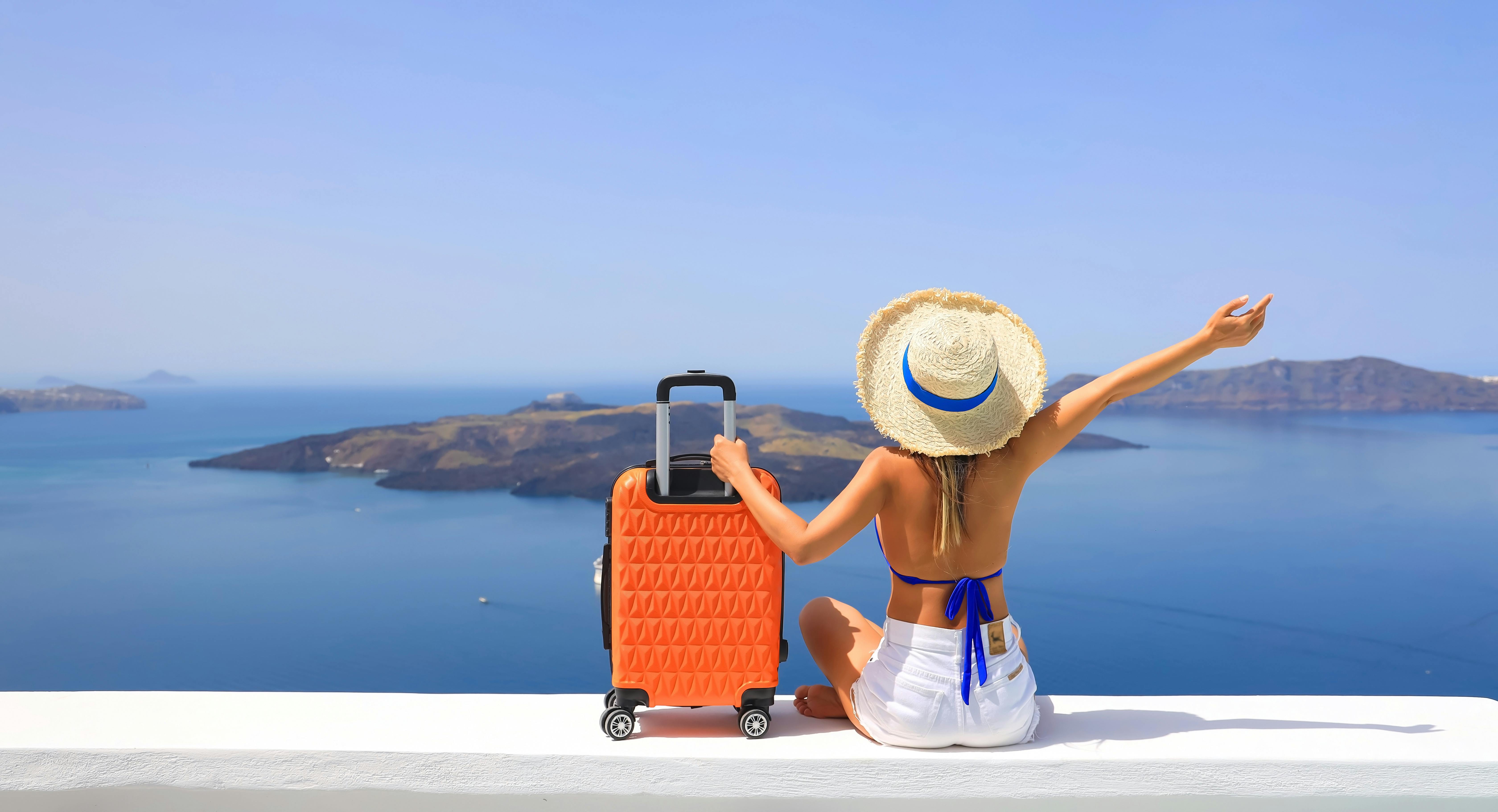 vrouw met koffer op de rand van een stenen muur kijkend naar de zee