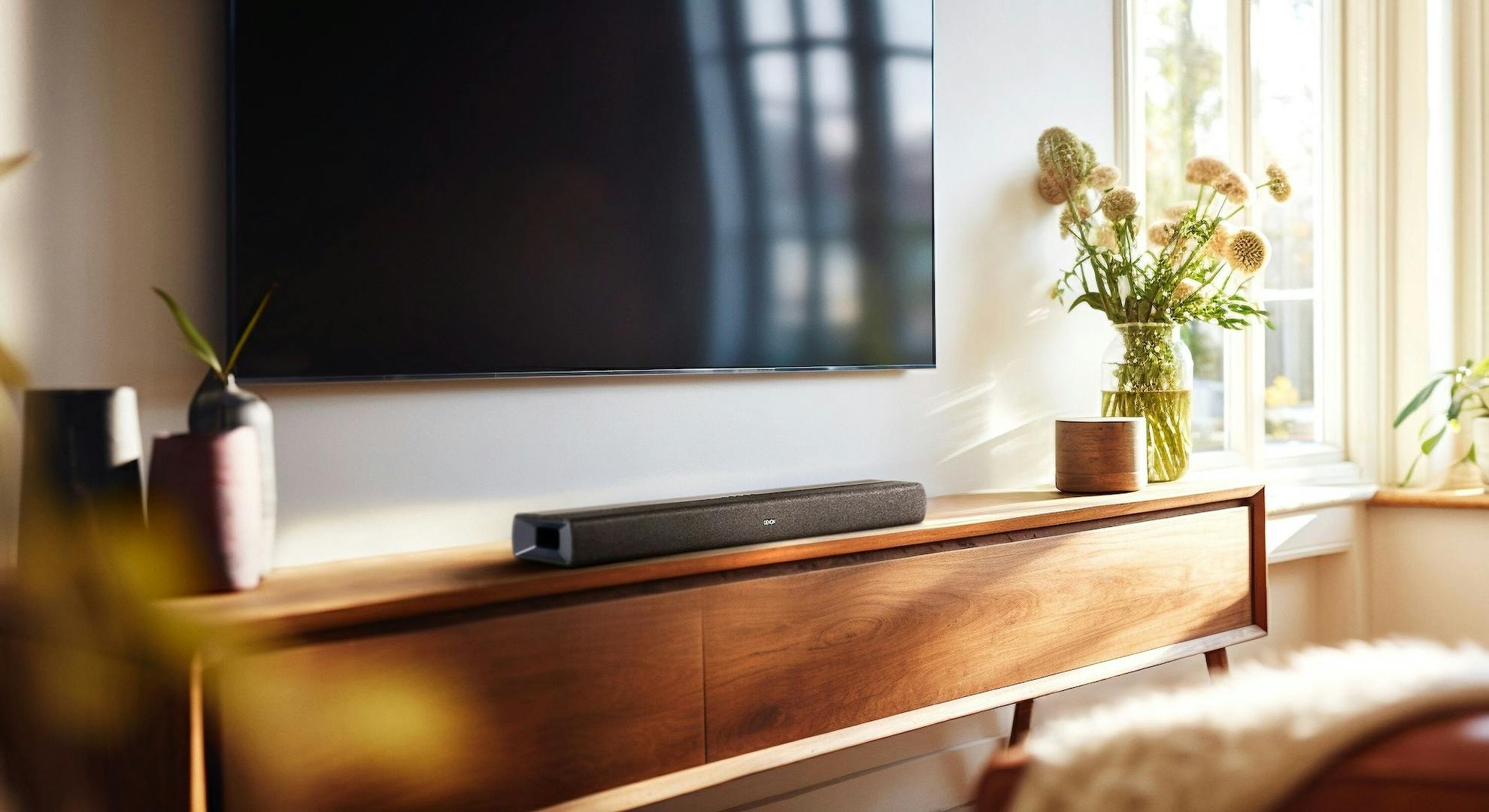 De Denon DHT-S218 op een kast bij een muurgemonteerde tv.