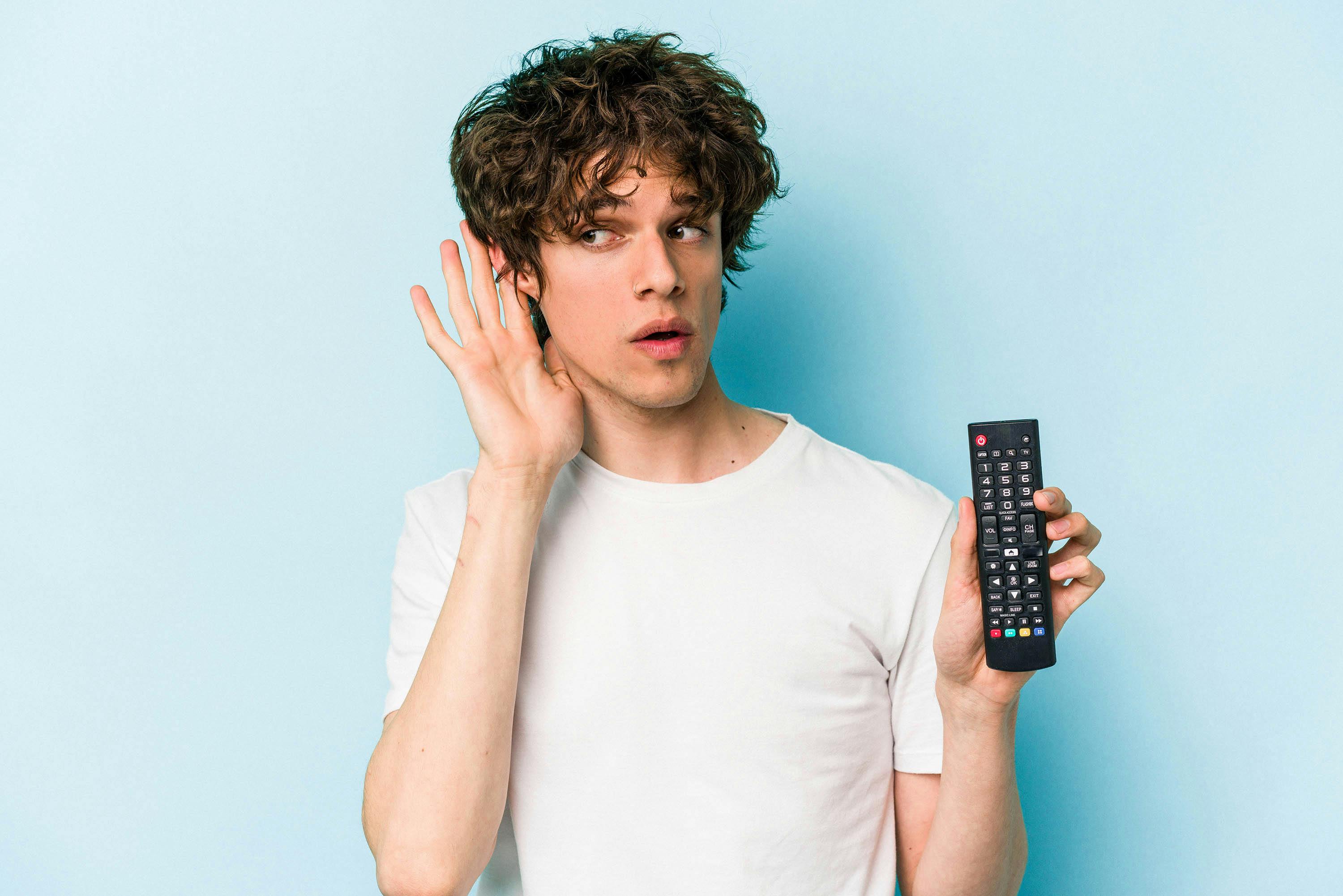 Man die een afstandsbediening van een televisie vasthoudt en zijn hand achter zijn oor houdt