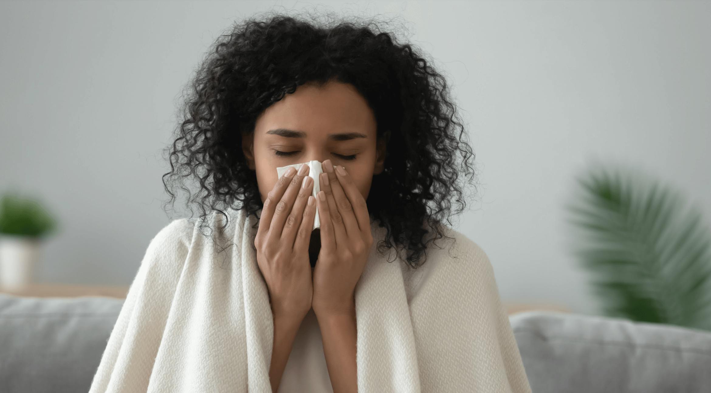 Woman blowing nose while sitting on couch with blanket over her