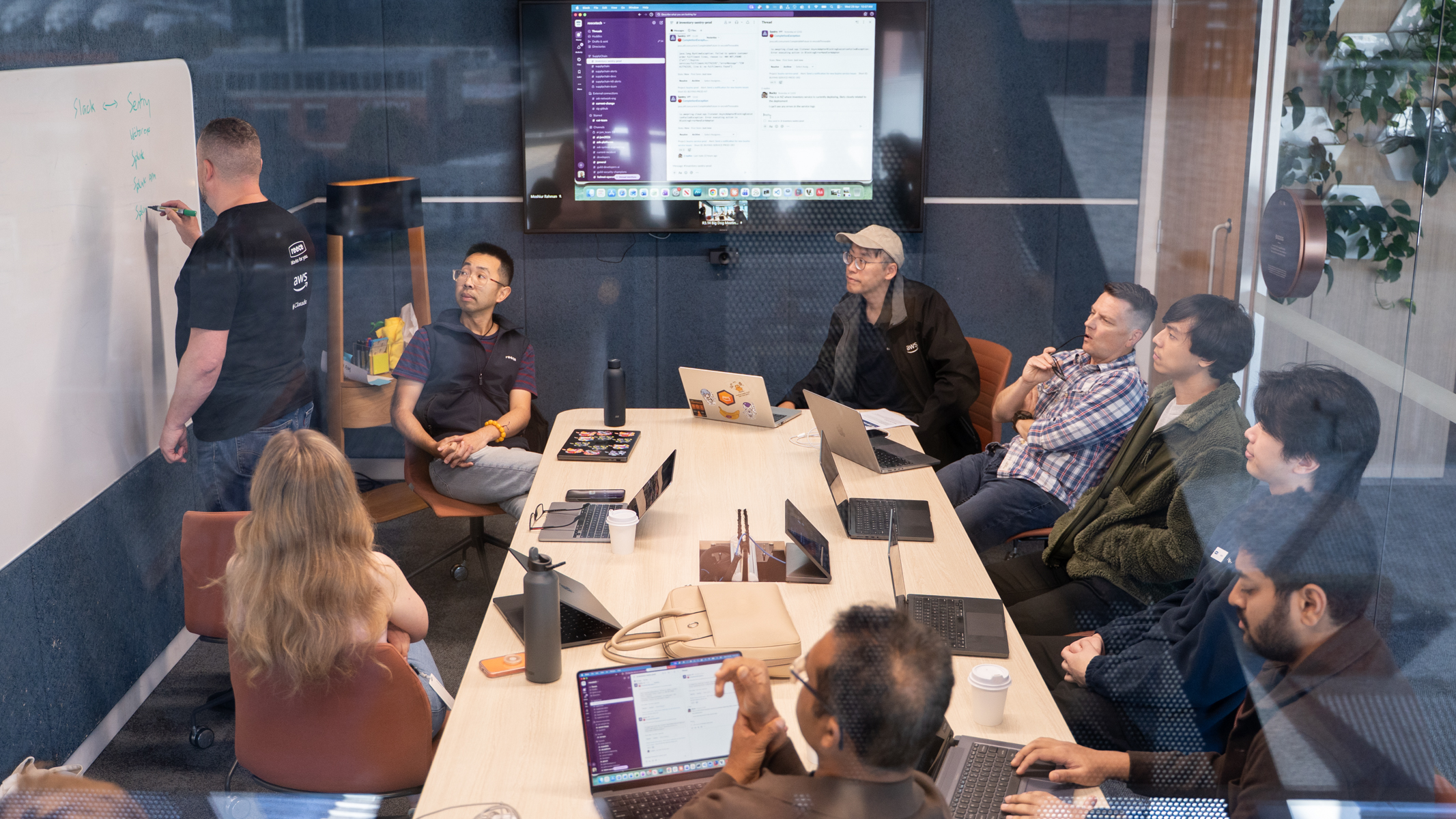 Team facing a whiteboard as a man writes on a board, nine people in the room, slack on the screen
