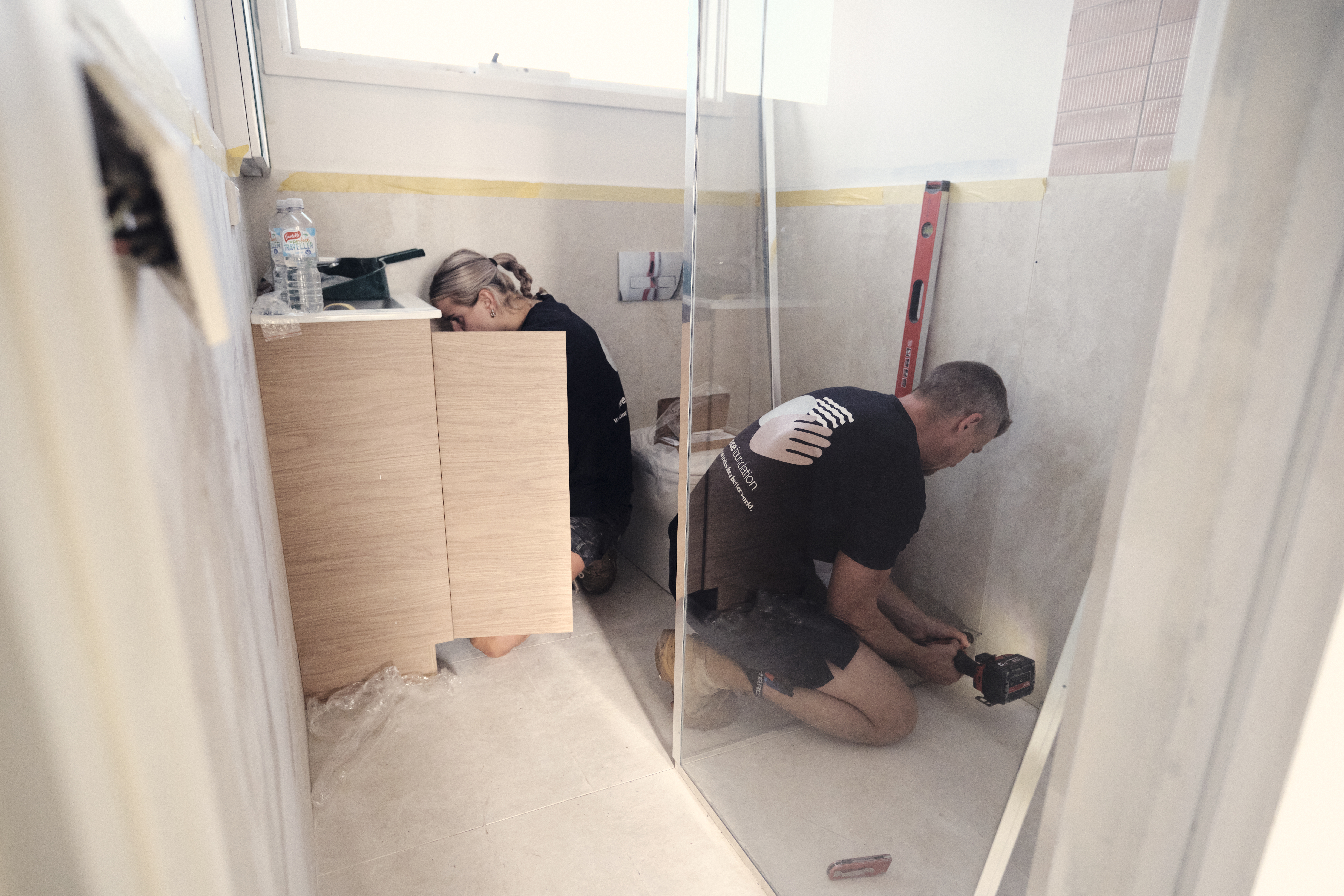 Two people working in a bathroom - On left, a woman finishes pluimbing under a cabinet, on the right, a man screws in a bracket for a glass shower wall.