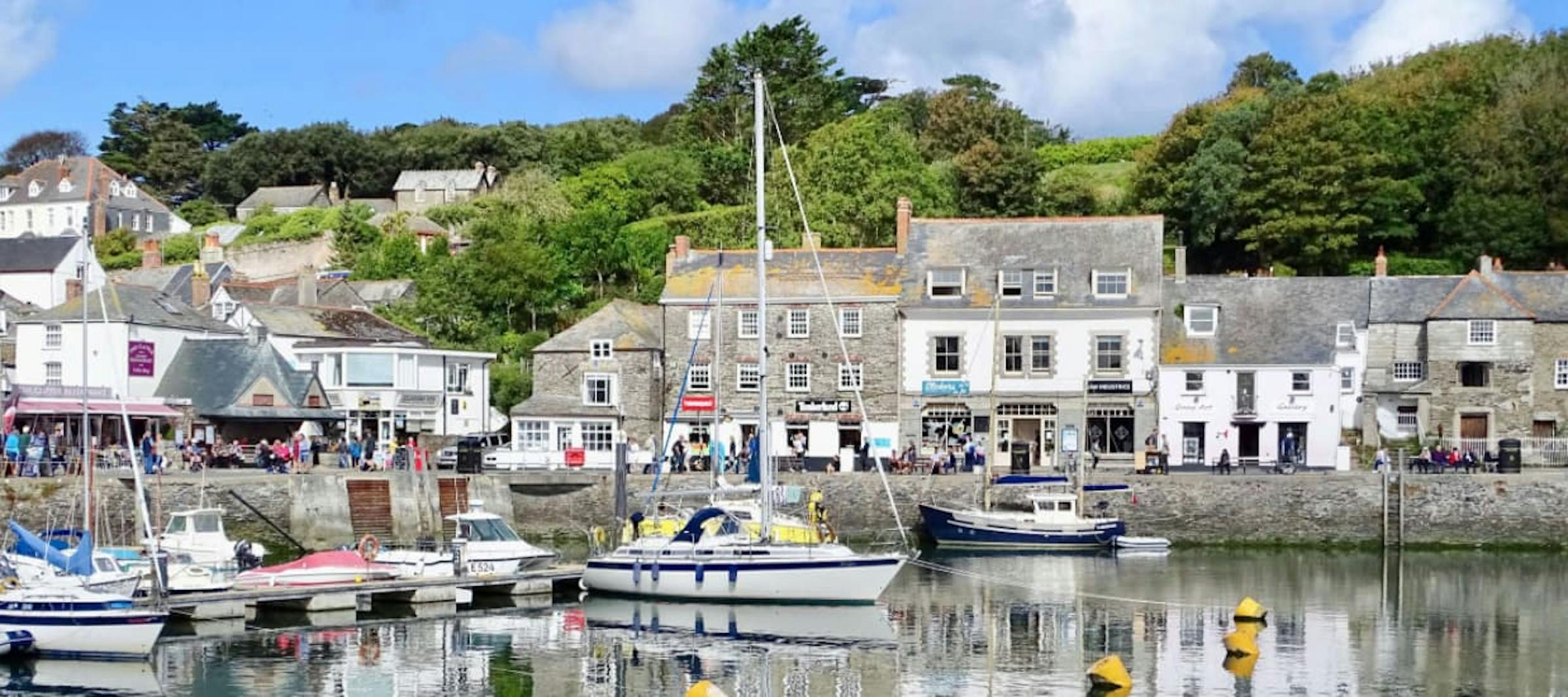 Sailboats fill Padstow harbour, framed by stone cottages and a green hillside in Cornwall.