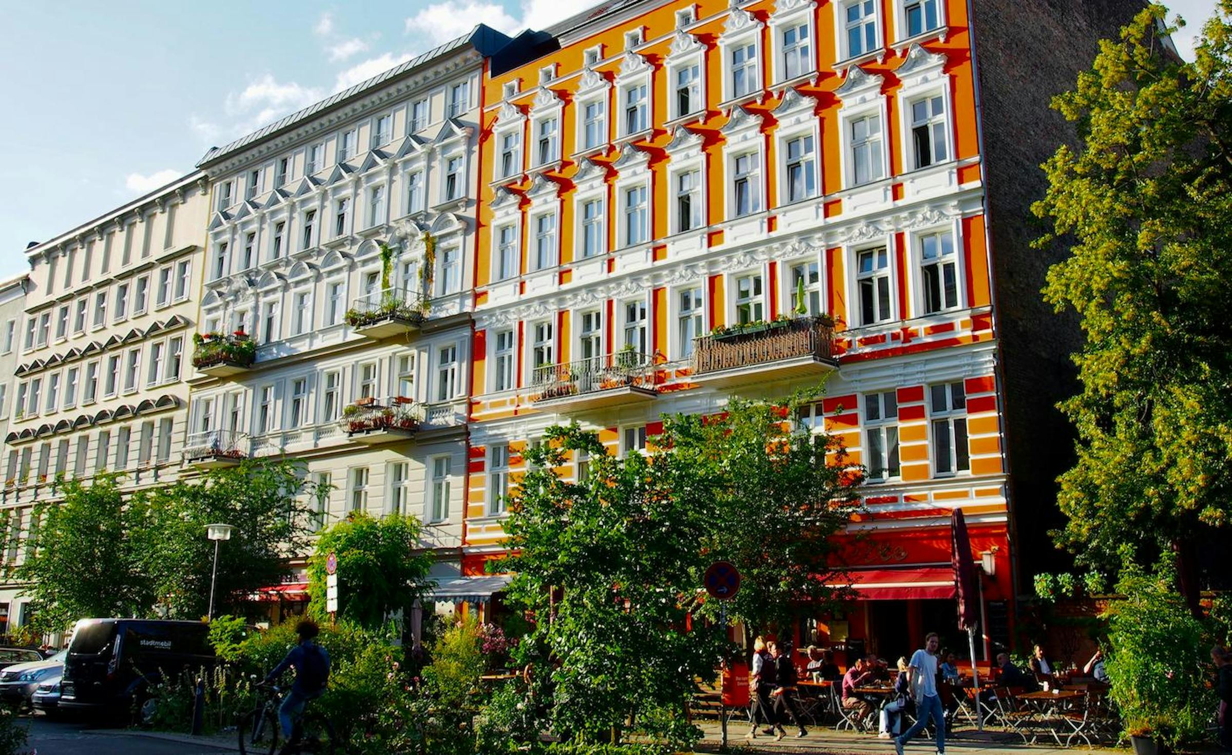 Ornate cream and orange apartment facades line a leafy Berlin street with a pavement café and cyclist.