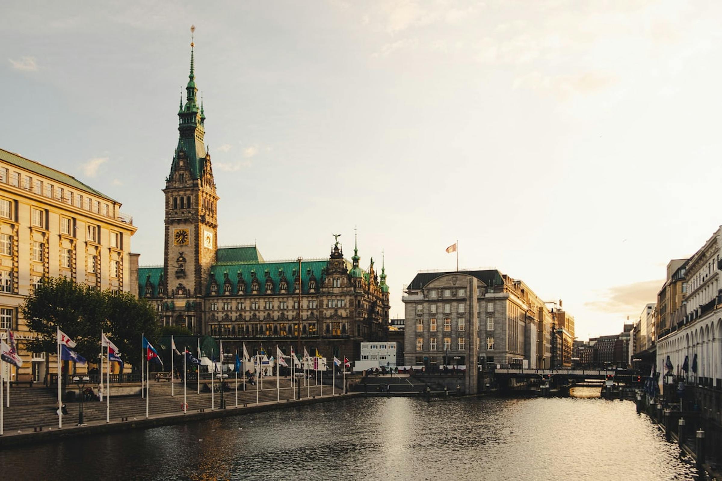 Hamburg's ornate Rathaus clock tower rises above a canal lined with flags at sunset.