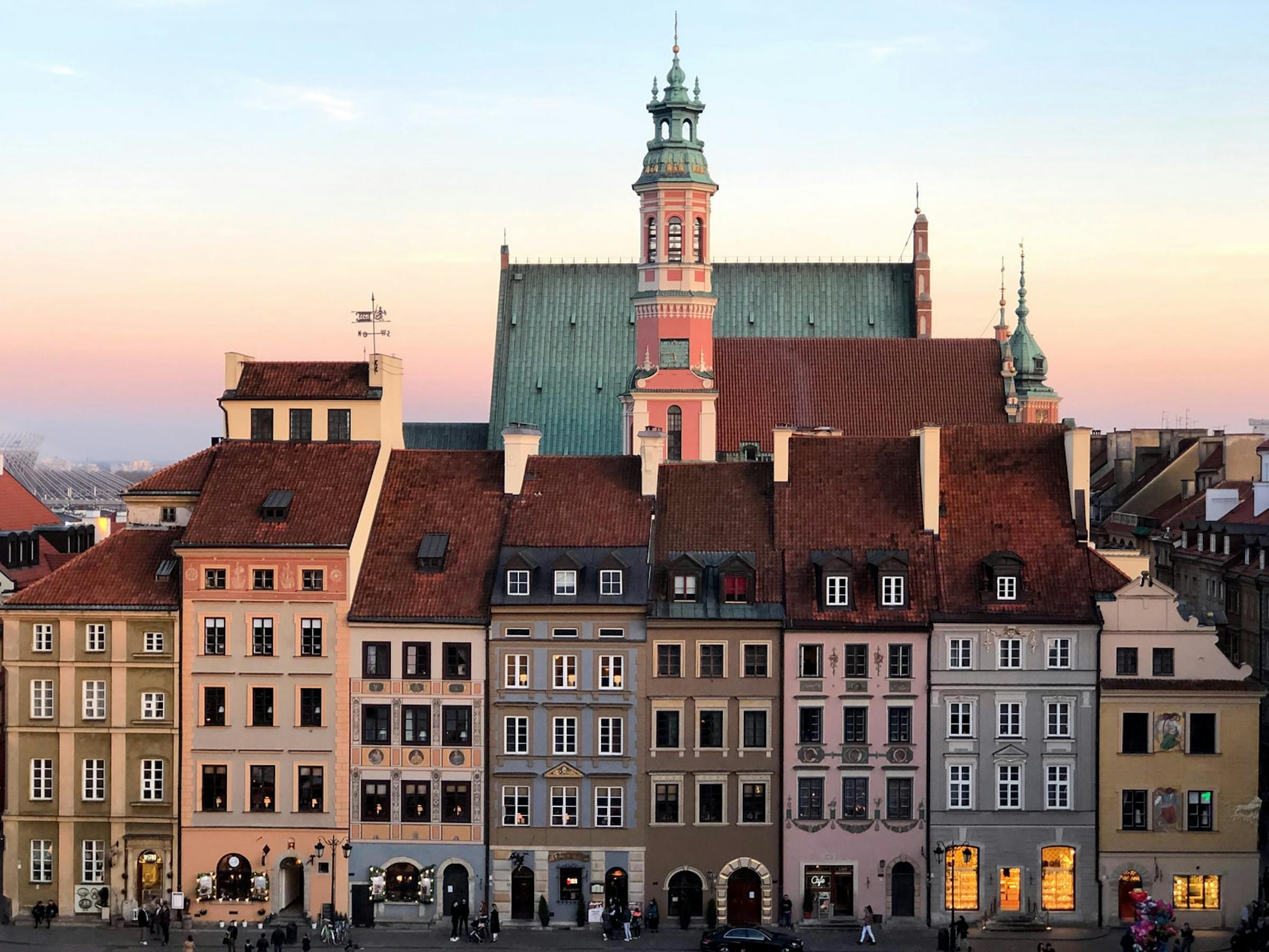Colourful townhouses line Warsaw's Old Town square beneath a domed church tower at sunset.