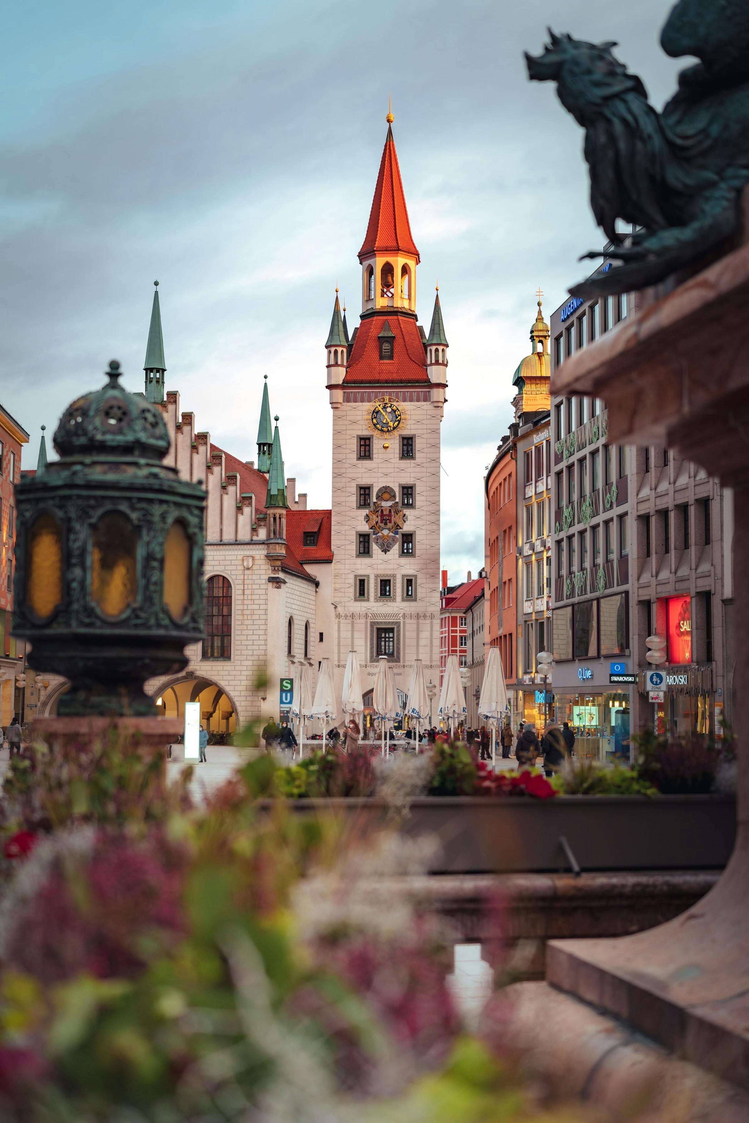The clock tower of Munich's Altes Rathaus rises above Marienplatz, framed by flowering plants in the foreground.