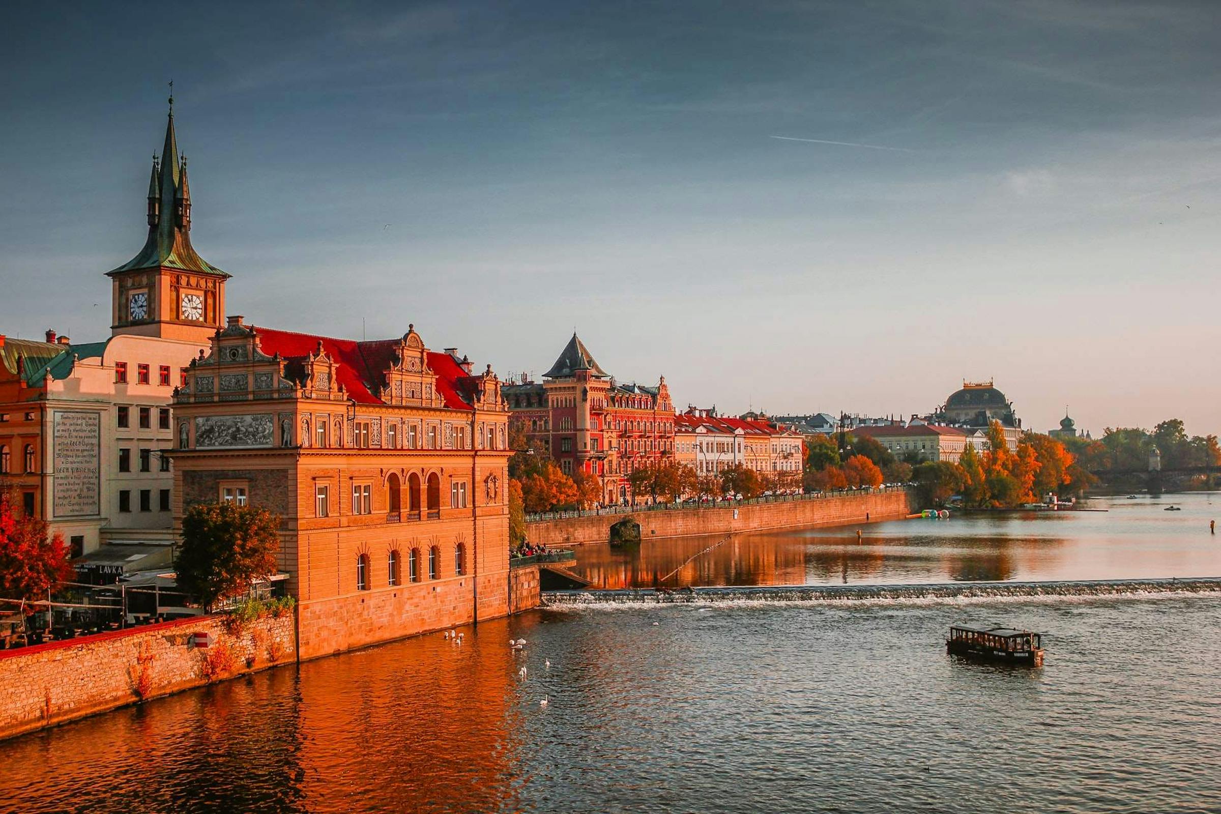 Golden-hour light bathes ornate riverside buildings along Prague's Vltava River in autumn.