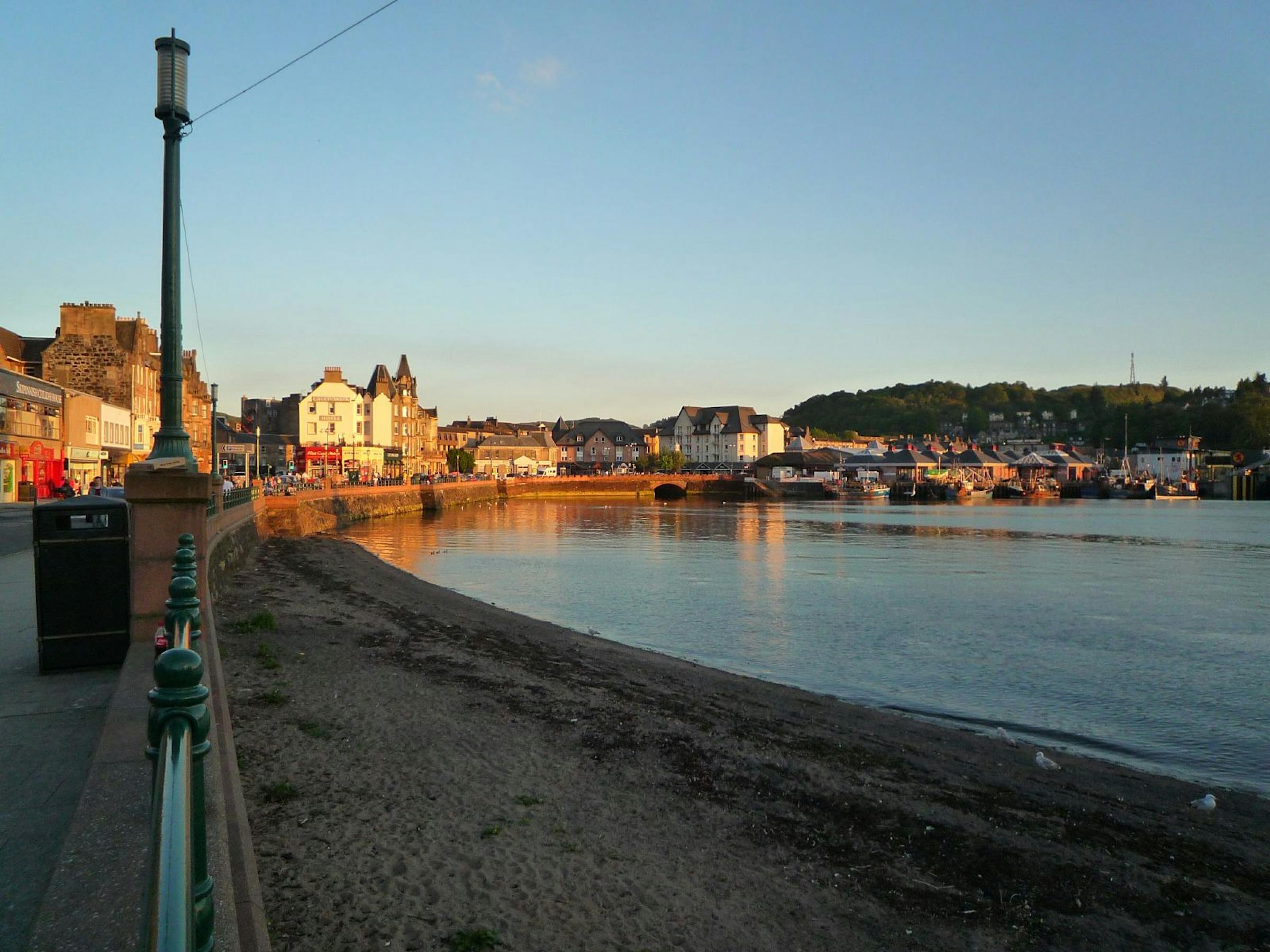 Oban's waterfront promenade and harbour buildings glow at sunset beside the calm bay.