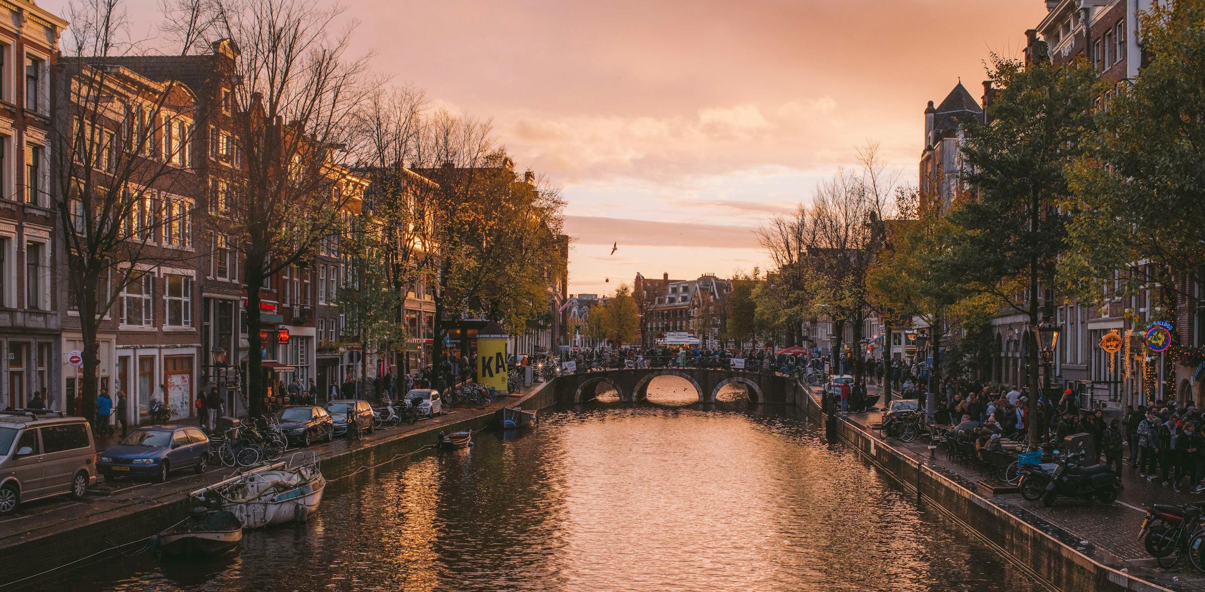 Amsterdam canal at golden-hour sunset, crossed by a stone arch bridge and lined with gabled houses and moored boats.