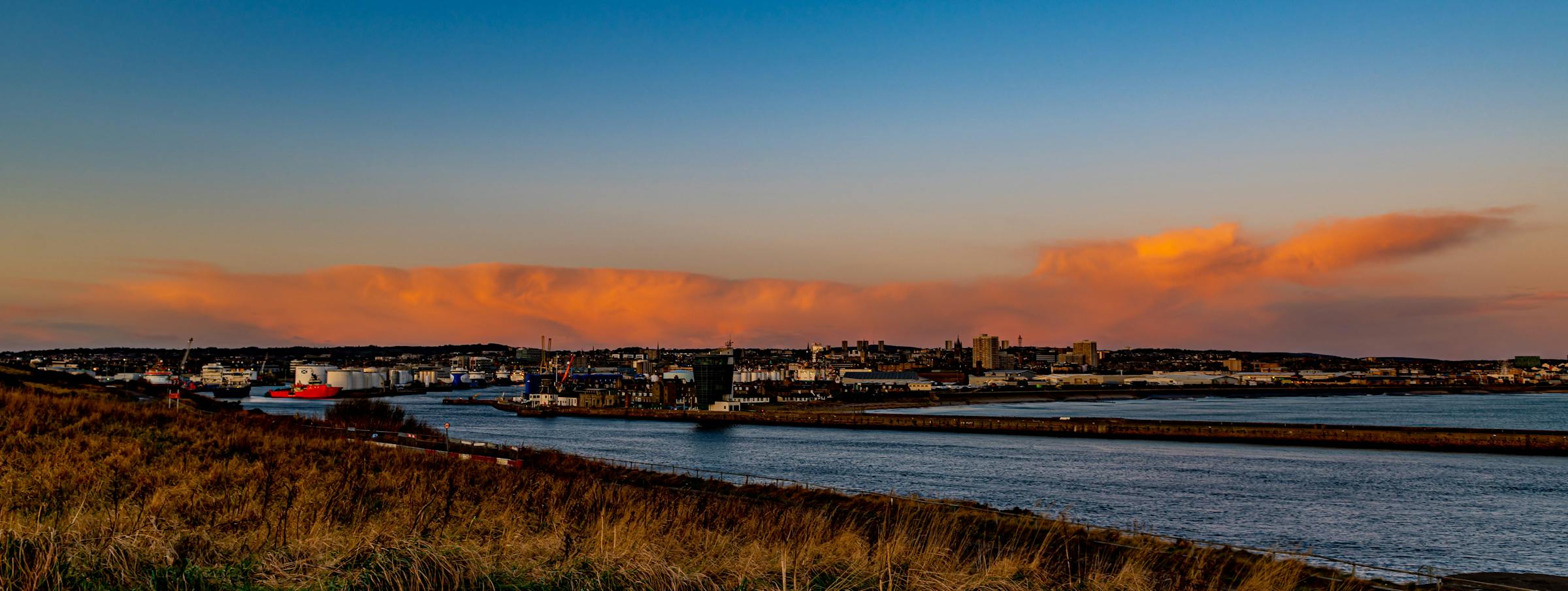 Aberdeen's harbour and skyline glow under sunset clouds, with a cargo ship docked at the quay.