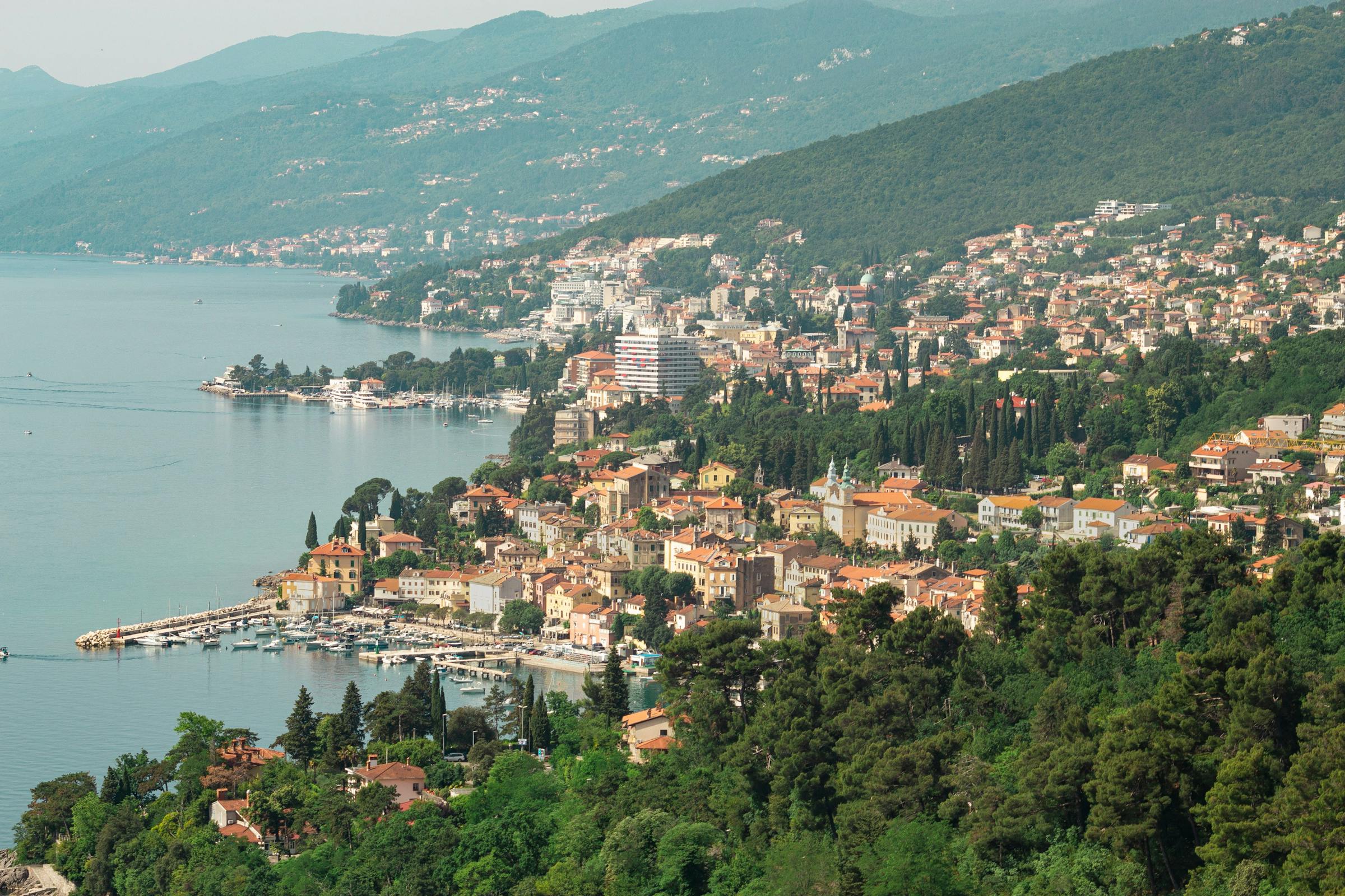 Terracotta rooftops of a Croatian coastal town spill down a forested hillside above a calm blue bay.