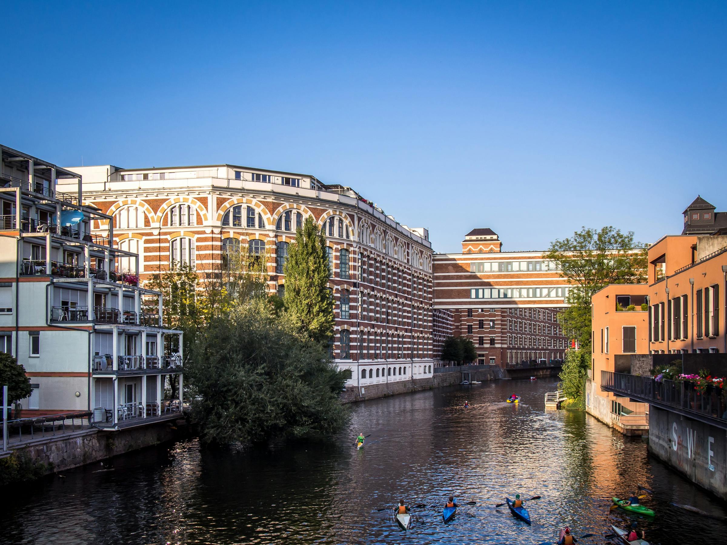 Kayakers paddle a canal past a converted red-brick industrial mill in Leipzig on a sunny evening.