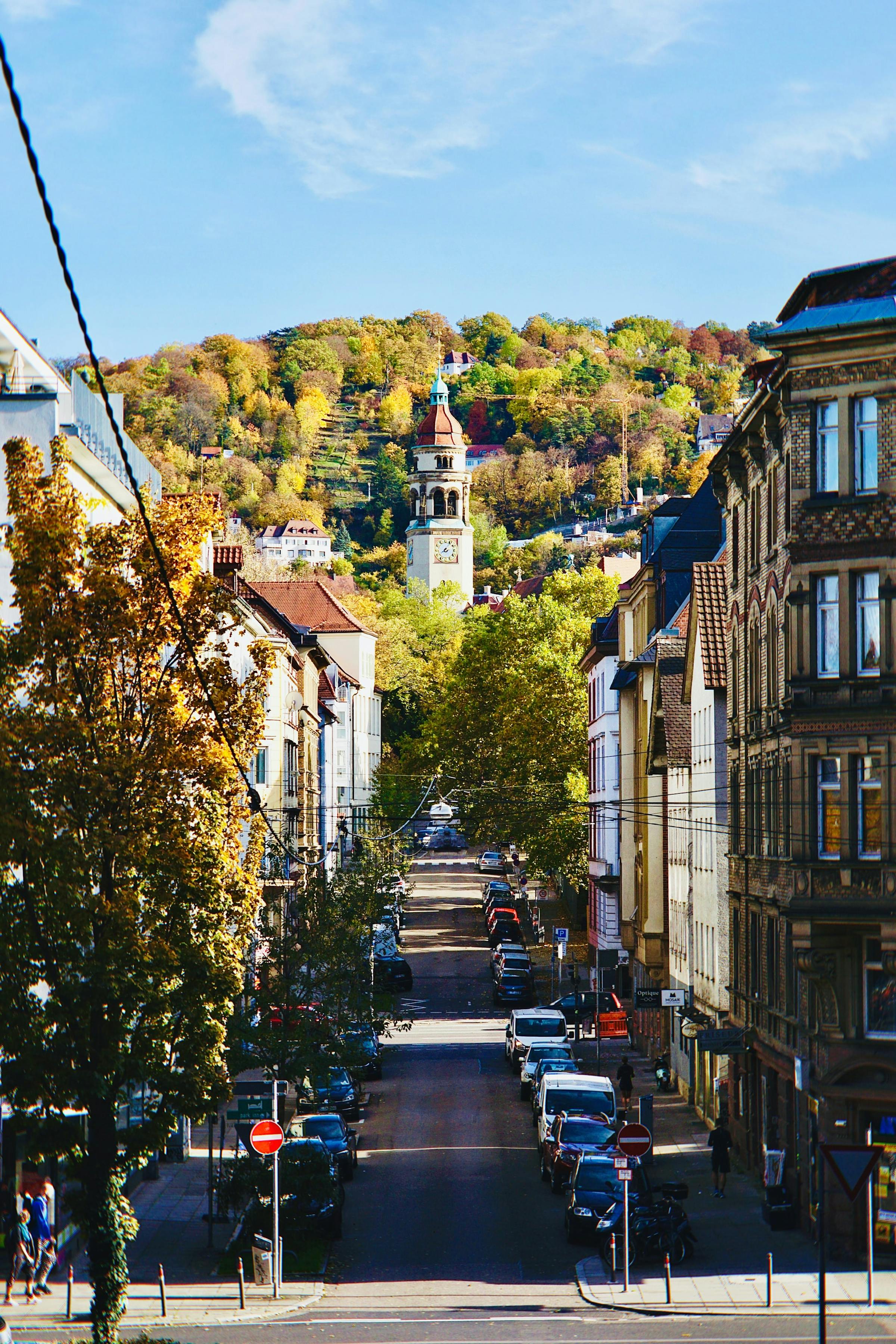 A tree-lined Stuttgart street rises toward a church tower on a wooded, autumn-coloured hillside.