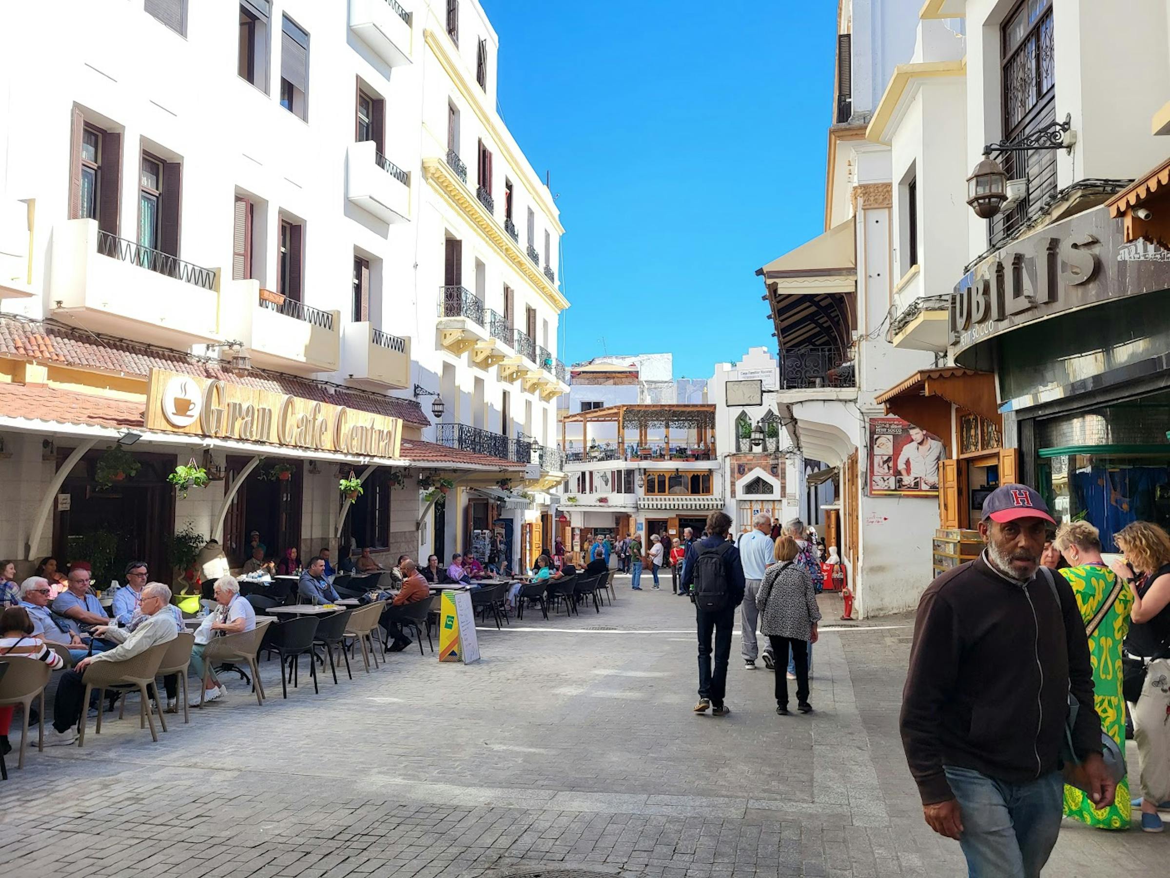 A sunny street in Tangier lined with a pavement café, whitewashed buildings, and passersby.