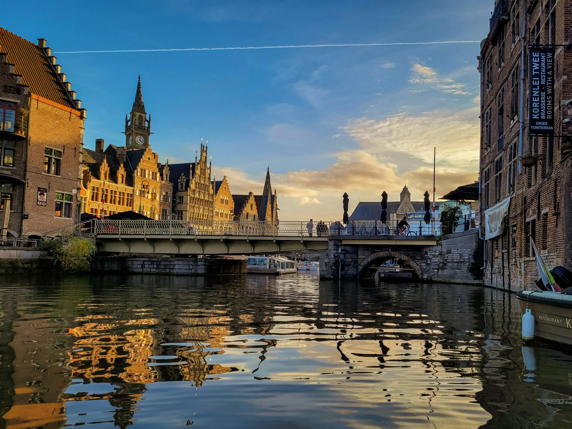 Ornate guild houses line Ghent's Graslei canal at sunset, with a stone bridge and moored boats.