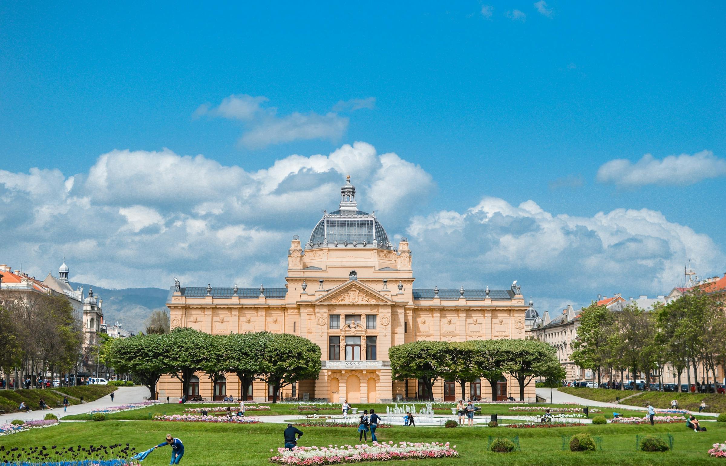 Zagreb's Art Pavilion, a domed yellow building, overlooks a park with a fountain and gardens.