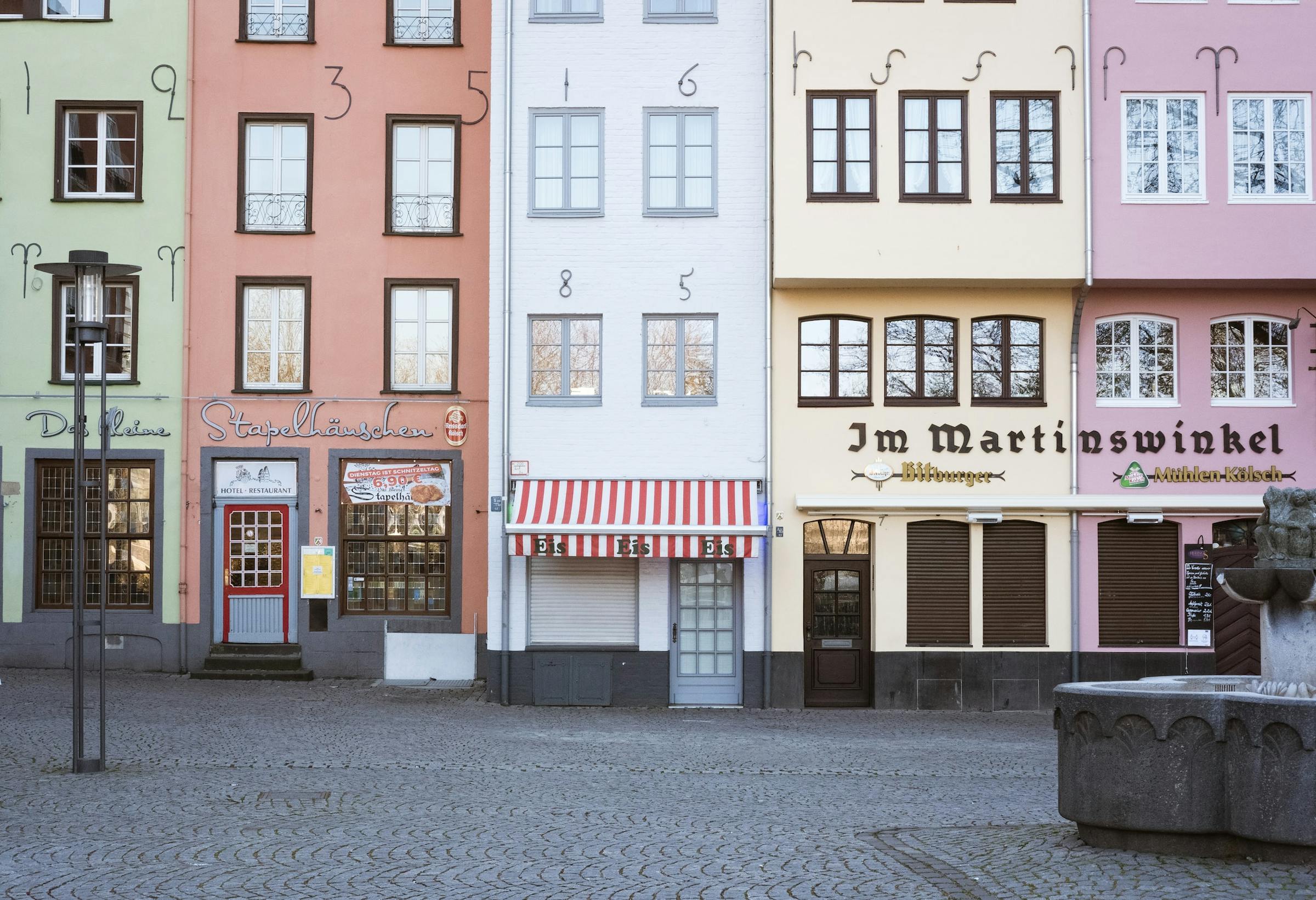 Pastel-painted terraced houses with a striped awning line a cobbled square in Cologne's old town.