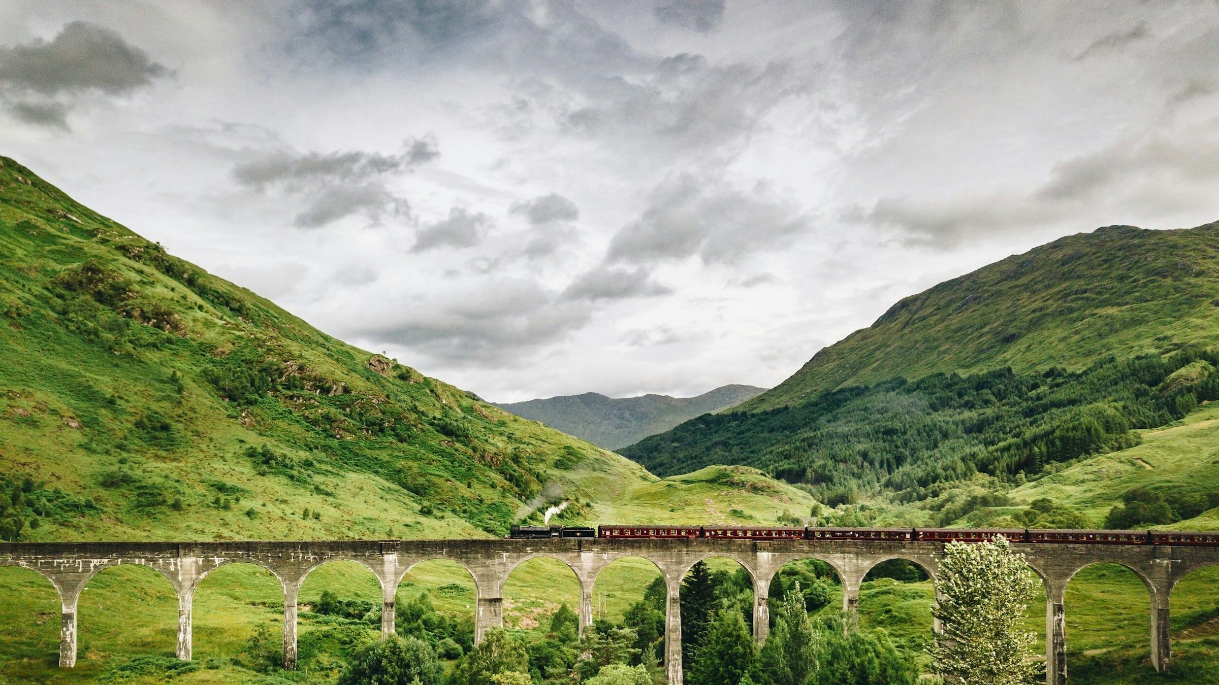A steam train crosses the curved Glenfinnan Viaduct through the green Scottish Highlands.